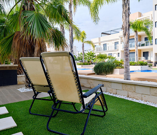 Two empty lounge chairs on artificial lawn grass inside of personal area backyard residential summer villa, view to modern homes with swimming pool, luxury, summer holidays lifestyle concept